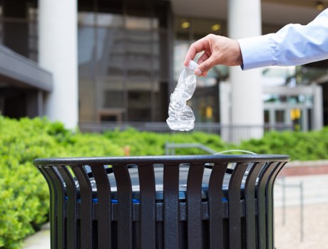 A person wearing a light blue long-sleeve shirt is disposing of a clear plastic bottle into a black outdoor trash bin with vertical slats, situated on a concrete pavement. The background features modern office or commercial building windows and a landscaped area with green bushes and plants, indicating an urban or business area possibly in Thamesmead. The person’s hand is visible holding the bottle above the open top of the bin, which is positioned in a paved outdoor setting suitable for rubbish collection. The scene emphasizes waste disposal activity related to commercial or office rubbish removal, aligning with services provided by Commercial Waste Thamesmead, with the environment well-maintained and clean, suggesting a focus on responsible waste management in the local area.