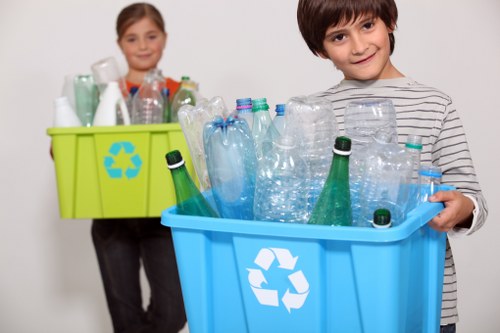 A young boy with dark hair, wearing a striped long-sleeve shirt, holds a blue recycling bin filled with empty plastic bottles of various shapes and sizes, predominantly clear with some green-tinted bottles. The bin features a white recycling symbol on its front and is placed on a flat surface indoors. In the background, a young girl with light brown hair, dressed in a light-colored top, stands holding a lime green recycling box filled with additional plastic bottles and containers. The setting appears to be a clean, neutral environment, likely a home or office space, emphasizing waste segregation and recycling practices typical of rubbish removal services in Thamesmead. The lighting is bright and even, highlighting the textures of the plastic bottles and the vivid colours of the containers. This image visually supports rubbish removal and recycling initiatives by [COMPANY_NAME] in the local Thamesmead area, illustrating the collection and sorting of recyclable plastic waste in a straightforward and professional manner.