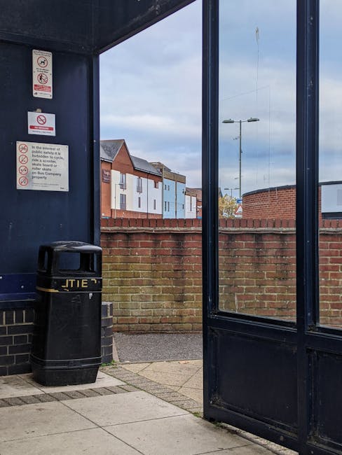 A small, semi-enclosed outdoor rubbish disposal area with dark blue painted metal walls and a glass door on the right side, leading to a paved sidewalk. Inside the enclosure, there is a black plastic 